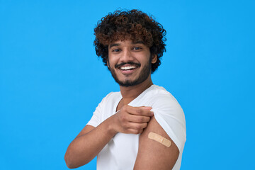 Happy young adult indian latin man showing bandage plaster on arm after getting vaccination isolated on blue. Vaccine and people inoculation, immunization for covid prevention concept. Portrait - obrazy, fototapety, plakaty