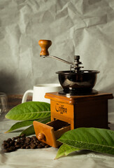 Coffee grinder with coffee beans and leaves falling nearby Set the scene for a photo with the mug and mug against a light gray background.