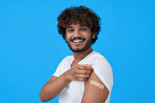 Happy young adult indian latin man showing bandage plaster on arm after getting vaccination isolated on blue. Vaccine and people inoculation, immunization for covid prevention concept. Portrait