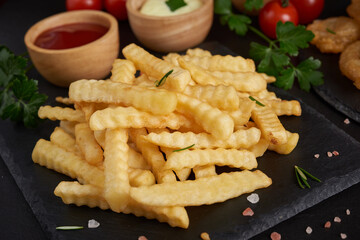 Homemade baked potato fries with mayonnaise, tomato sauce and rosemary on paper over black stone background. . tasty french fries, unhealthy food.