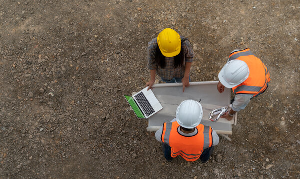  team of engineers and co-workers wearing safety uniforms and engineer helmets holding construction blueprints, checking construction exactly as they were working on a housing project's laptop. 