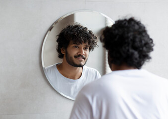 Handsome indian guy looking at his reflection in the mirror, standing in modern bathroom at home