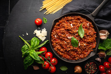 classic italian bolognese sauce stewed in a pan with ingredients on black tile background, top view