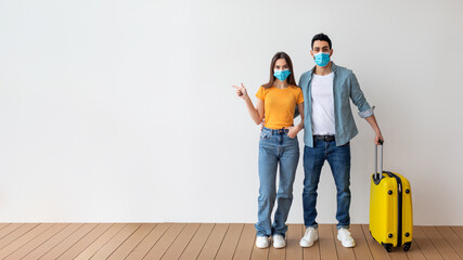 Young spouses wearing medical face masks standing with suitcase, woman pointing aside at free space