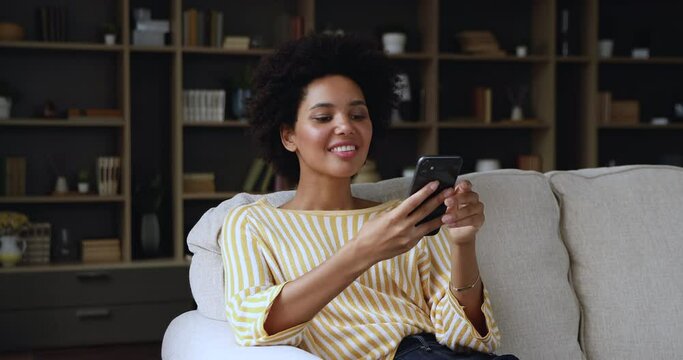 Pretty African woman sit on sofa use smartphone, share messages, chatting with boyfriend in social media enjoy free time at home. Modern wireless tech usage, connection, virtual communication concept