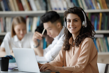 Happy gen Z Latin student girl in headphones working on research study project in library, watching online presentation on laptop, learning webinar, sitting by classmates. Head shot portrait