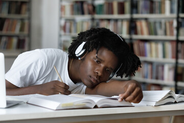 Tired bored lazy Black student listening to audio lesson, webinar, writing notes, reading open book...