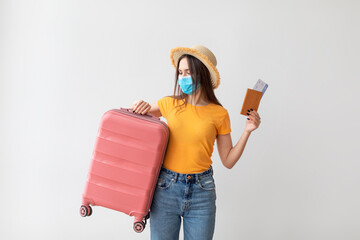 Tourism during COVID-19 pandemic. Young woman wearing medical face mask, holding suitcase and passport
