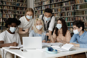 Obraz premium Interracial group of gen Z students wearing facial medical masks sitting at laptop in college library, watching online presentation, discussing learning webinar, working on project together