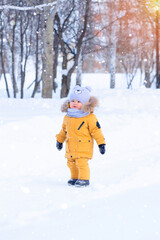 Fototapeta na wymiar A portrait of a toddler 15-20 months old in yellow warm clothes stands on a path in the middle of a winter park during a snowfall