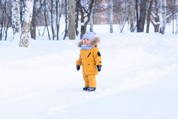 Fototapeta na wymiar Portrait of a toddler 15-20 months old in yellow warm clothes in a winter park looks at the falling snowflakes with surprise
