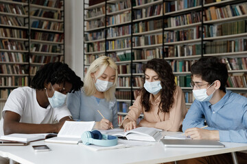 Diverse college students in protective medical masks writing notes, summary in public library, studying in group together, working on learning project, reading book, textbook, talking