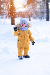 Fototapeta na wymiar The kid greets by raising his hand and looking forward. Portrait of a toddler 15-20 months old in yellow warm clothes during a snowfall.