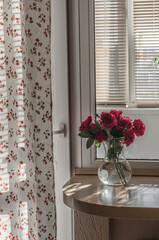 A bouquet of garden roses in a vase with water on a window sill against the background of a window and a loggia, a curtain in a small flower.