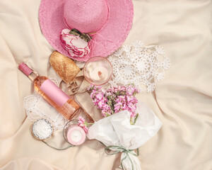 Picnic on beige blanket: pink sun hat, rose wine bottle with empty label, wine glass, baguette, cheese, candles and pink flowers bunch. Romantic outdoor lunch idea with food and drink. Top view