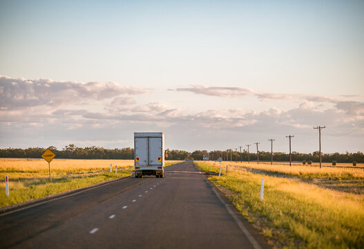 A truck travels along an empty long straight road in the New South Wales countryside on a blue sky day at sunset.