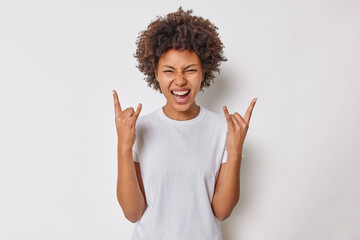 Carefree joyful woman with curly hair makess rock n roll gesture heavy metal sign enjoys favorite...