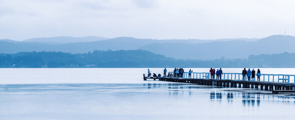 People walking along a long wooden jetty on a calm lake.  Long Jetty, New South Wales, Australia.