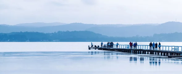 Fotobehang Pier People walking along a long wooden jetty on a calm lake.  Long Jetty, New South Wales, Australia.  © Scott Donkin
