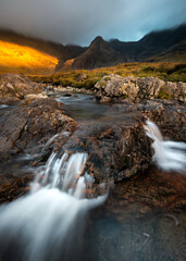 Fairy Pools Waterfall with view of famous Cuillin mountain range. Beautiful golden evening light with dark and dramatic clouds. Isle of Skye, Scotland, UK.