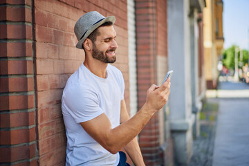 Handsome man using smartphone in the city smiling, leaning against the wall