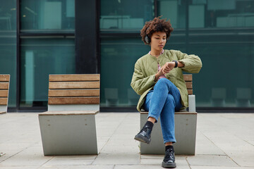 Full length shot of fashionable woman poses at bench waits for public transport checks time on wearable wristwatch listens music via headphones spends time in city reads message on smartwatch