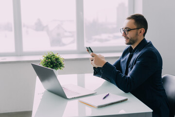 Business man sitting at desk in his office, holding phone and smiling happily while reading funny...