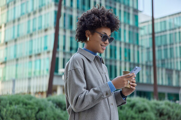 Horizontal shot of cheerful curly haired woman wears trendy sungasses and jacket holds mobile phone...