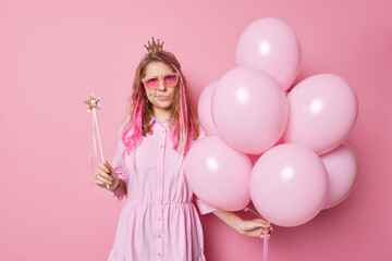 Obraz na płótnie Canvas Displeased irritated birthday girl has image of princess wears crown sunglasses and dress holds magic wand and bunch of balloons being on party isolated over pink background. Celebration concept