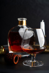 Obraz na płótnie Canvas A glass of cognac and a pipe for tobacco against the background of a bottle of cognac and a candle on a dark surface