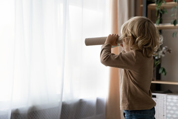 Side back view happy adorable little curly blonde kid boy looking in carton spyglasses, imagining...