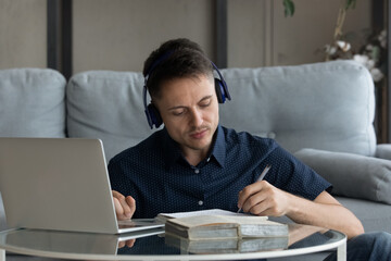 Focused young man wearing wireless headphones, listening educational lecture on computer, making notes in copybook improving knowledge, preparing for examination at home, e-learning concept.