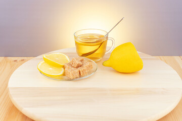 Tea in a glass cup with a spoon, half a lemon, pieces of brown sugar with lemon slices on a wooden table. 