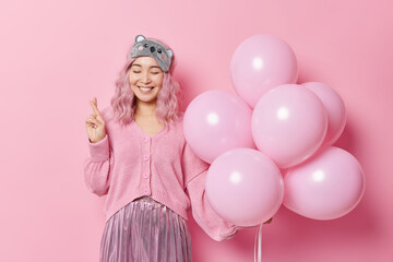 Obraz na płótnie Canvas Positive hopeful woman with dyed hair smiles happily keeps fingers crossed believes in good luck holds bunch of balloons celebrates birthday isolated over pink background believes dreams come true