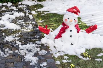 Unhappy snowman in mittens, red scarf and cap is melting  outdoors in sunlight on snowy green grass with small yellow flowers near wet pavement. Approaching spring, warm winter, climate change concept