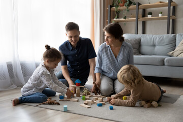 Happy bonding young couple parents and joyful little preschool children son daughter playing toys, sitting on floor carpet, enjoying creative playtime hobby activity together in modern living room.