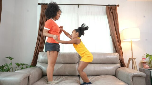 African family spending time together at home. Two little girl sibling dancing and jumping on sofa in living room with happiness. Happy child girl kid enjoy and fun indoor lifestyle playing together