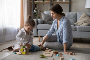 Adorable small preteen kid girl playing toys with affectionate mother or caring babysitter, sitting together on floor carpet at home. Loving young single mum having fun with preschool child daughter.