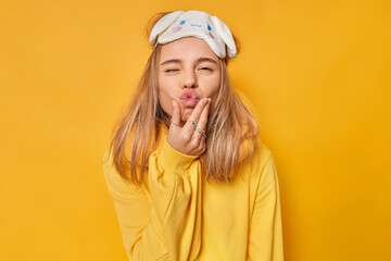 Funny sleepy woman keeps lips rounded wears blindfold neckpillow and casual jumper wants to kiss you poses against vivd yellow background. People rest sleeping and facial expressions concept