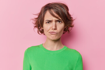 Fototapeta premium Portrait of young displeased woman frowns face has sulking expression purses lips has short hairstyle wears green jumper isolated dover pink studio background. Negative human emotions concept