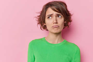 Frustrated young brunette woman purses lips raises eyebrows looks away feels displeased wears green jumper feels unhappy isolated over pink background blank copy space for your promotional content