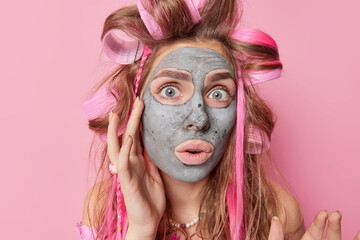 Portrait of surprised young woman applies moisturizing clay mask on face for skin treatment undergoes anti aging procedures wears hair curlers looks impressed at camera isolated over rosy wall