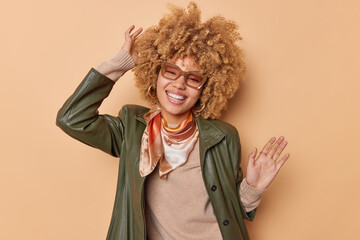 Relaxed happy woman with curly bushy fair hair stands over beige background dances happily to music lifts arms dressed in fashionable clothes smiles broadly feels free and carefree enjoys life