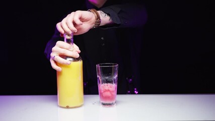 A bartender pours orange juice from a bottle into a glass glass glass with pink ice on a dark background, a white table