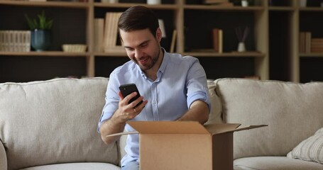 Millennial man sit on sofa at home opens parcel box, use phone check tracking number, quality of ordered goods and date, feels satisfied. Client enjoy webshop services and e retail app usage concept