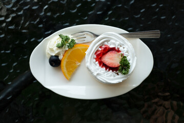 strawberry shortcake decorate on white dish with blue berry, whiped cream and orange tkae from top view, black background
