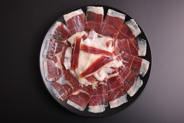 Portion of acorn-fed Iberian ham 100% Dehesa de Extremadura on a black plate and black background decorated with acorns