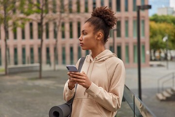 Curly sportswoman has break on training uses smartphone for blogging focused away thoughtfully sends text messages wears sweatshirt walks against urban background gets sportive notification.