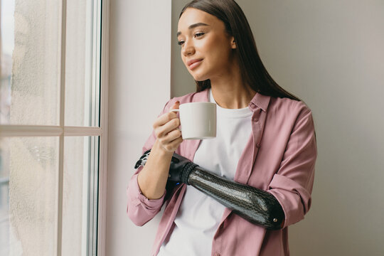 Picture of pretty attractive cyber woman with artificial iron robotic arm of black color standing next to window, looking through on city streets, holding white cup with hot drink in hand