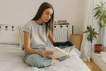 Smart intelligent woman reading sci-fi book flipping it through with metal fingers of her artificial black iron hand prosthesis sitting on bed in front of window with domestic plants on windowsill